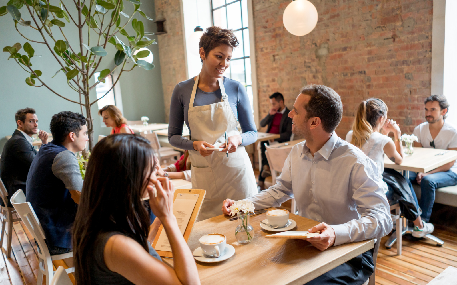 Tables at a restaurant with two people sitting at a table and a server smiling while taking their order. 