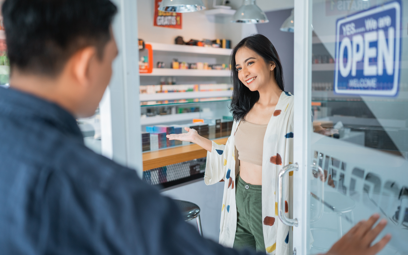 A woman opening the door to a vape store smiling at a customer. 