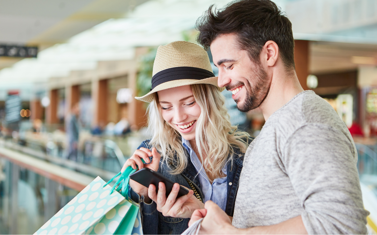 A man and a woman smiling and looking at phone while the woman holds shopping bags